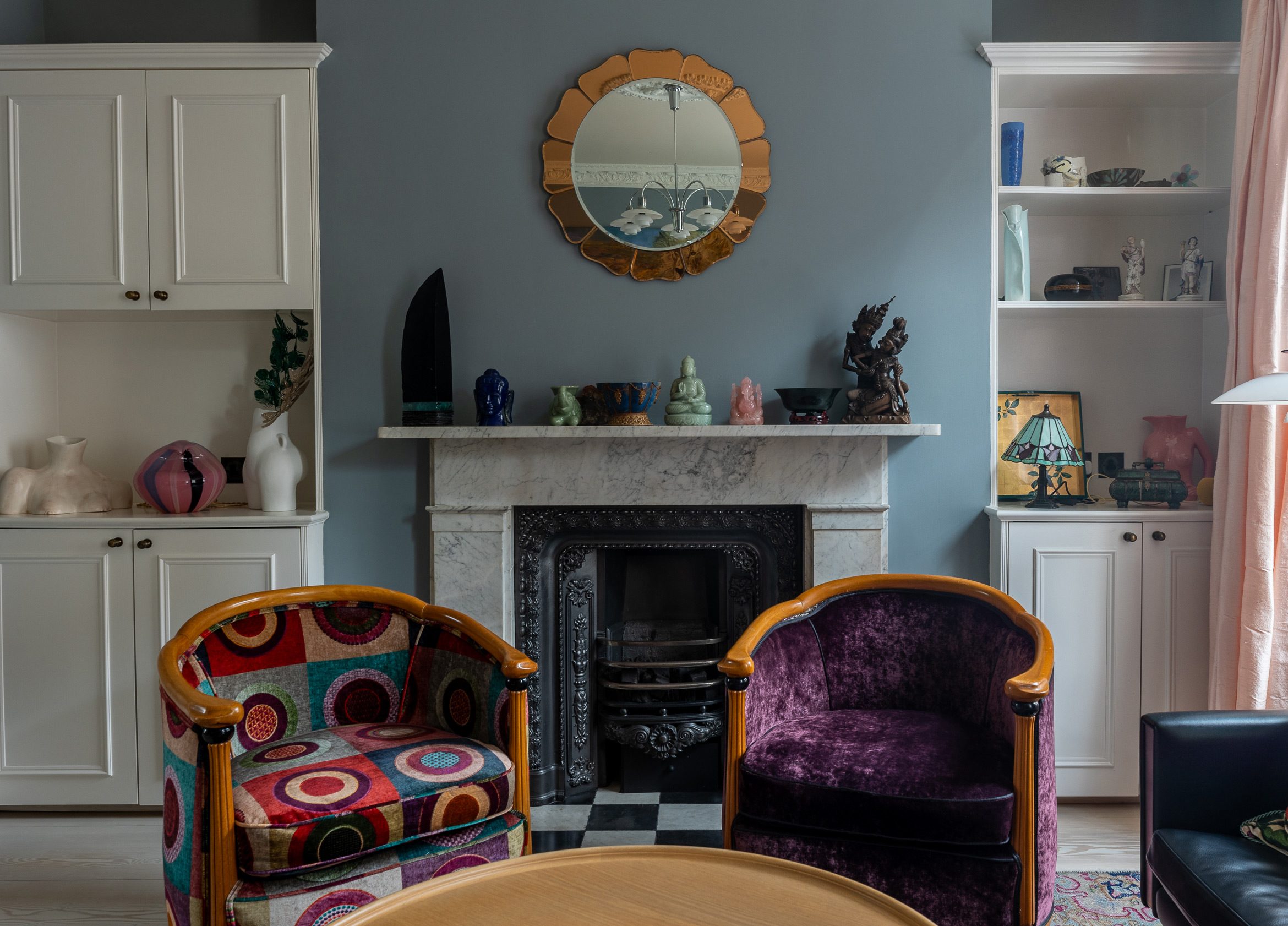 A cozy living room with a marble fireplace as the focal point. Two colorful armchairs sit in front of it. A round mirror hangs above the mantel, flanked by white shelving units displaying various decorative items.