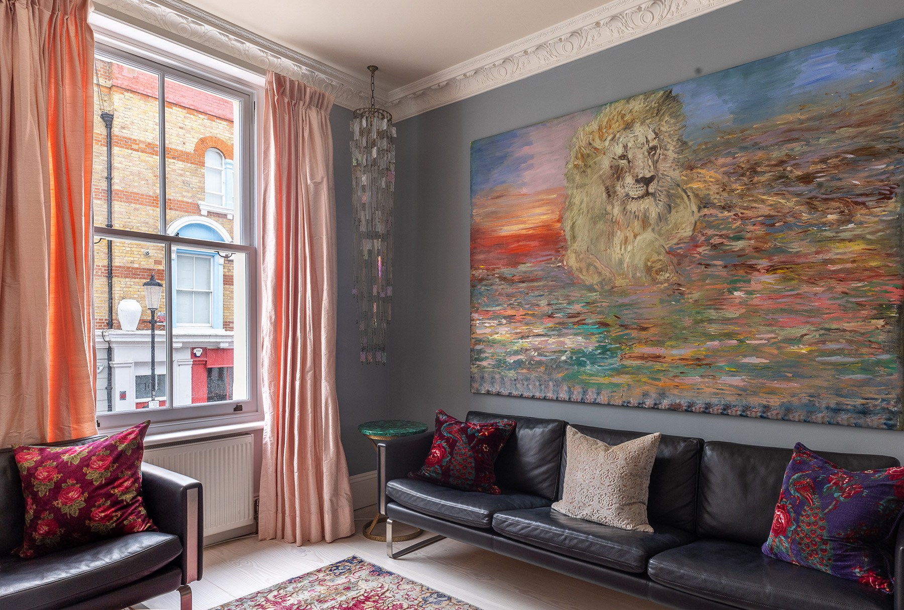 A cozy living room features a large lion painting on the wall, a black leather sofa with colorful cushions, and a tall stained glass lamp. Pink curtains frame a window with a view of a brick building across the street. A small round table sits beside the sofa.
