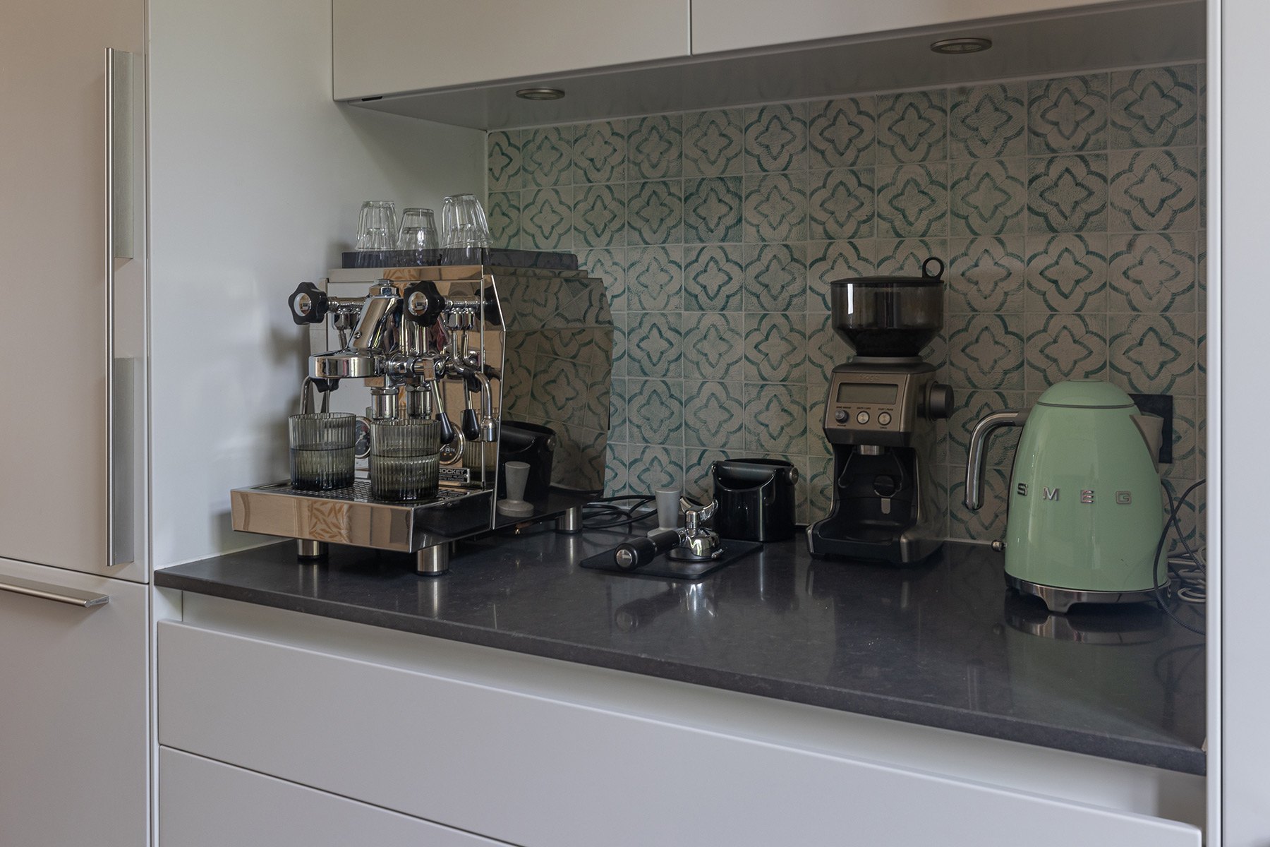 Modern kitchen counter with a silver espresso machine, a coffee grinder, and a green electric kettle. The backsplash features a pattern of blue and white tiles. The cabinets are white, and the countertop is dark gray.