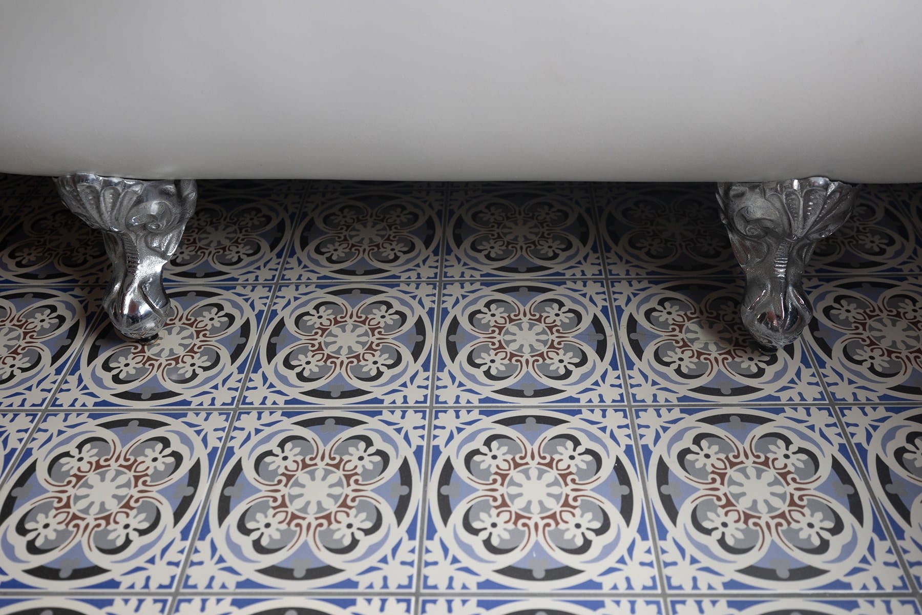 A close-up of an ornate tiled floor with a vintage clawfoot bathtub. The floor features a repeating pattern of blue, white, and brown circular designs. The clawfoot is metallic and intricately detailed.