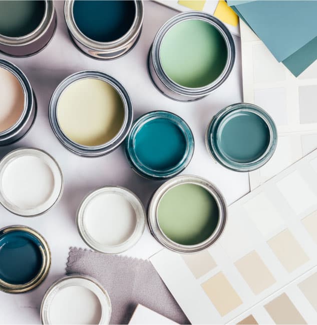 Top view of several open paint cans displaying a variety of colors, including greens, blues, and whites. Surrounding the cans are paint sample cards and fabric swatches, suggesting a color selection or interior design project.