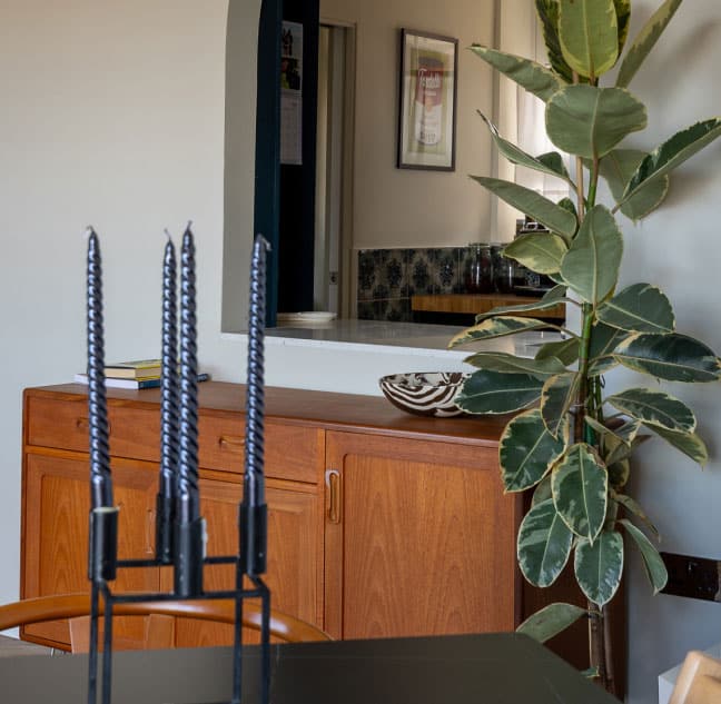 A modern interior with a wooden sideboard, a tall leafy plant, black twisted candles in a holder, and a decorative bowl. A framed picture and a glimpse of a tiled wall are in the background.