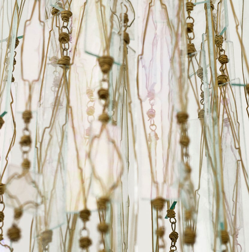 Close-up of a decorative hanging installation featuring delicate strings, translucent sheets, and knotted cords creating an intricate pattern with light shining through, casting subtle shadows and highlights.