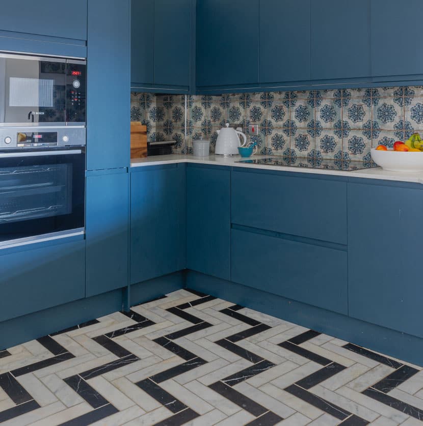 A modern kitchen with blue cabinets and a patterned backsplash featuring blue and white floral tiles. The oven is built into the cabinetry. The floor has a herringbone pattern in black and white. A bowl of fruit sits on the countertop.