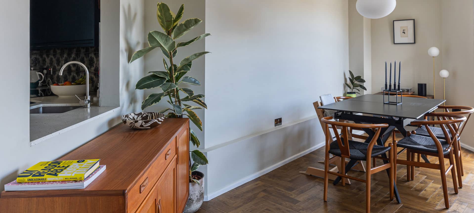 A dining room with a dark rectangular table surrounded by wooden chairs. A large plant stands near the white wall. A sideboard holds a yellow book, and a kitchen area is visible through an open hatch. A white pendant light hangs from the ceiling.