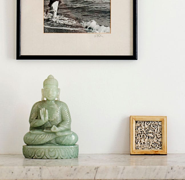 A jade Buddha statue is placed on a marble surface. Above it hangs a framed black-and-white photograph of a person by the sea. Next to the statue, theres a small ornate square object with a detailed pattern.