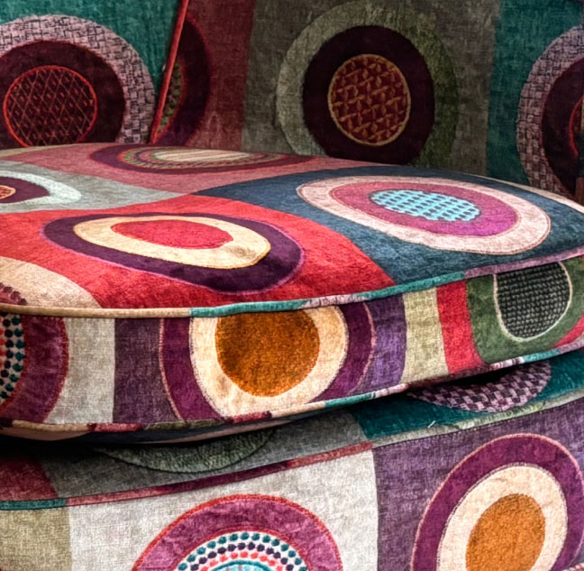 Close-up of a fabric chair with a vibrant circular pattern in various colors, including red, purple, blue, and green. The overlapping circles have textured designs, giving the fabric a dynamic and lively appearance.