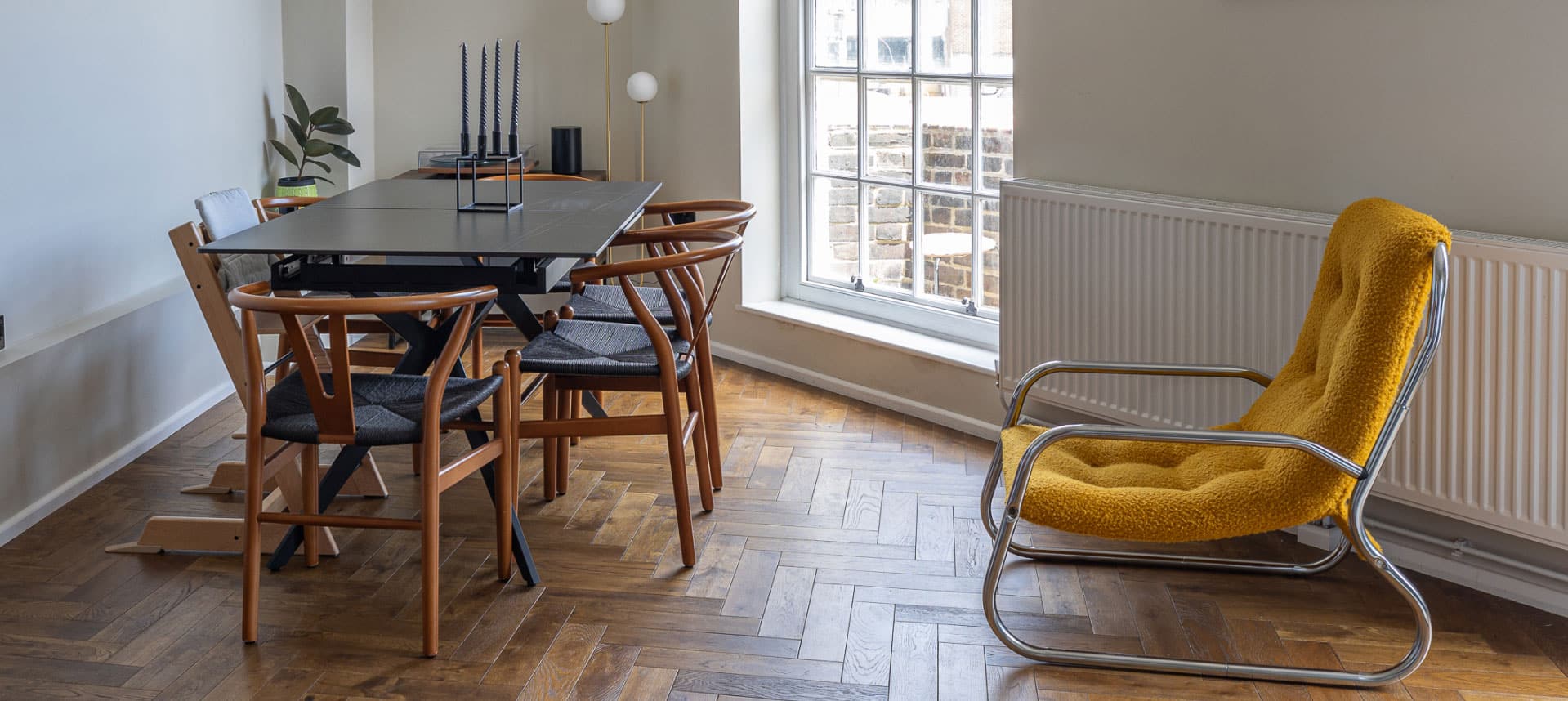 A modern dining room with a black rectangular table and four wooden chairs. A mustard-yellow cushioned chair sits nearby. The room features a large window, parquet flooring, and neutral-colored walls with a radiator below the window.
