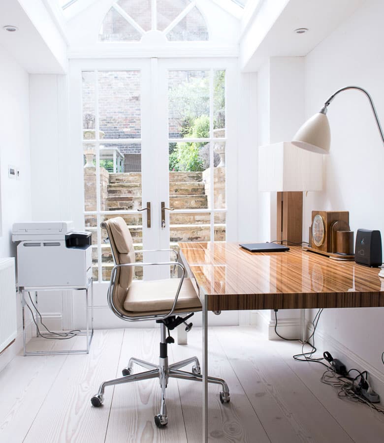 A bright home office with a wooden desk and a swivel chair. A laptop and lamp sit on the desk. Large glass doors behind the desk lead to stone steps and a garden. A printer is on the left side, adding functionality to the sleek setup.