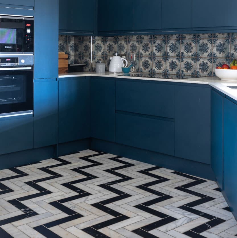 A modern kitchen features dark blue cabinets, patterned blue and white backsplash tiles, and a herringbone black-and-white tiled floor. A white kettle and fruit bowl rest on the countertop.