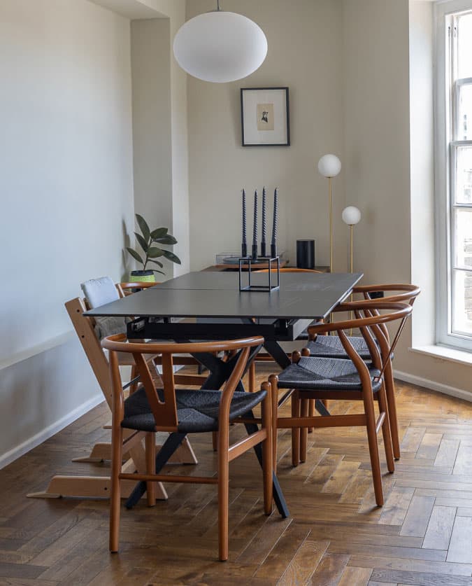 A modern dining area with a black rectangular table and six wooden chairs with dark cushions. A round white ceiling lamp hangs above. A small plant and minimalist decor are on a side ledge. Daylight streams through a tall window onto the herringbone floor.