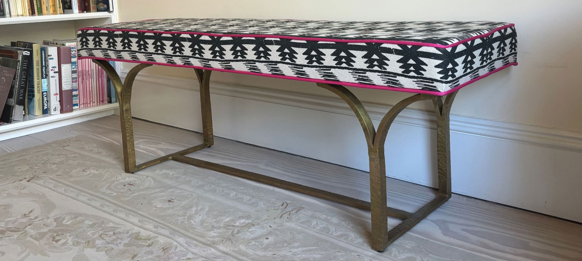 A cushioned bench with a geometric black and white pattern and pink trim on four curved bronze legs. Its placed on a patterned rug next to a white bookshelf filled with books.