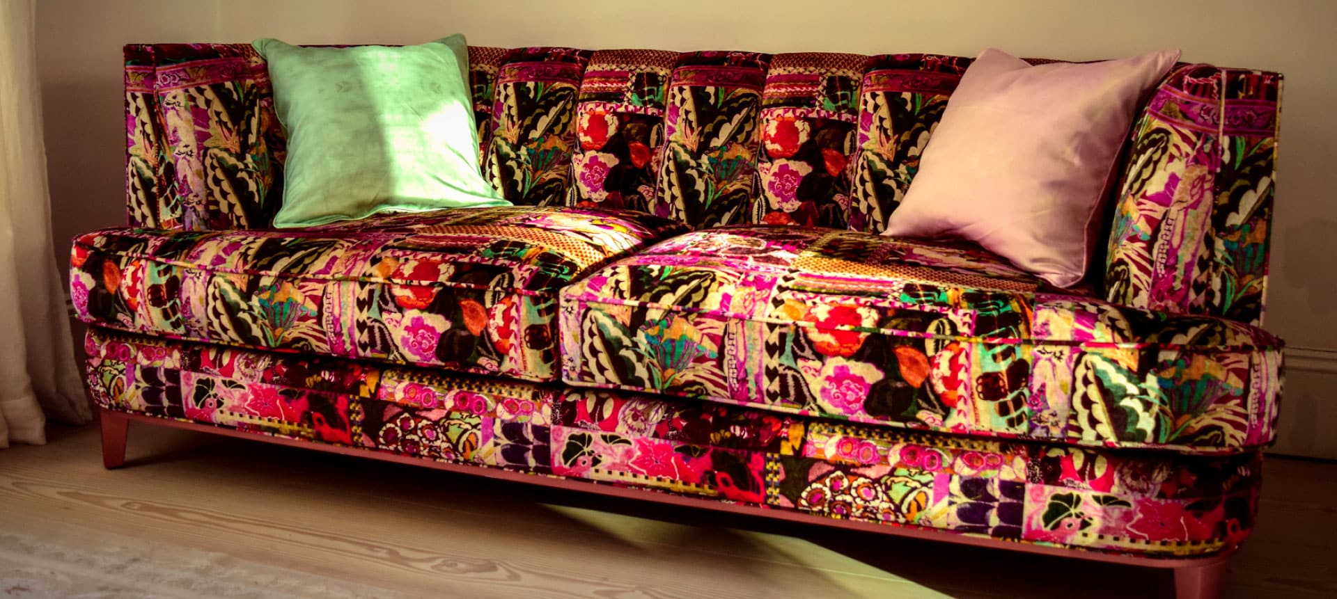 A colorful, floral-patterned sofa with wooden legs is positioned against a light-colored wall. Two pastel pillows, one green and one pink, are placed on the sofa. Sunlight casts a warm glow on the vibrant fabric.