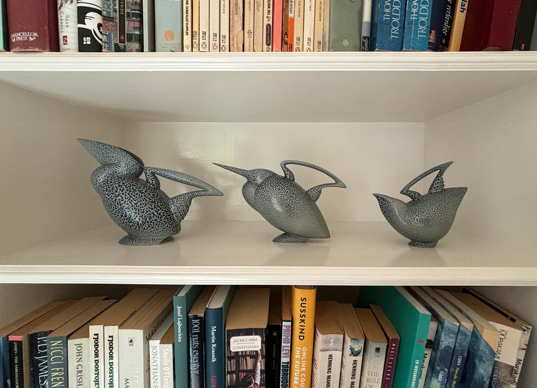 Three bird sculptures with intricate patterns are displayed on a white shelf. The shelf is surrounded by books arranged vertically and horizontally, featuring various titles and authors.