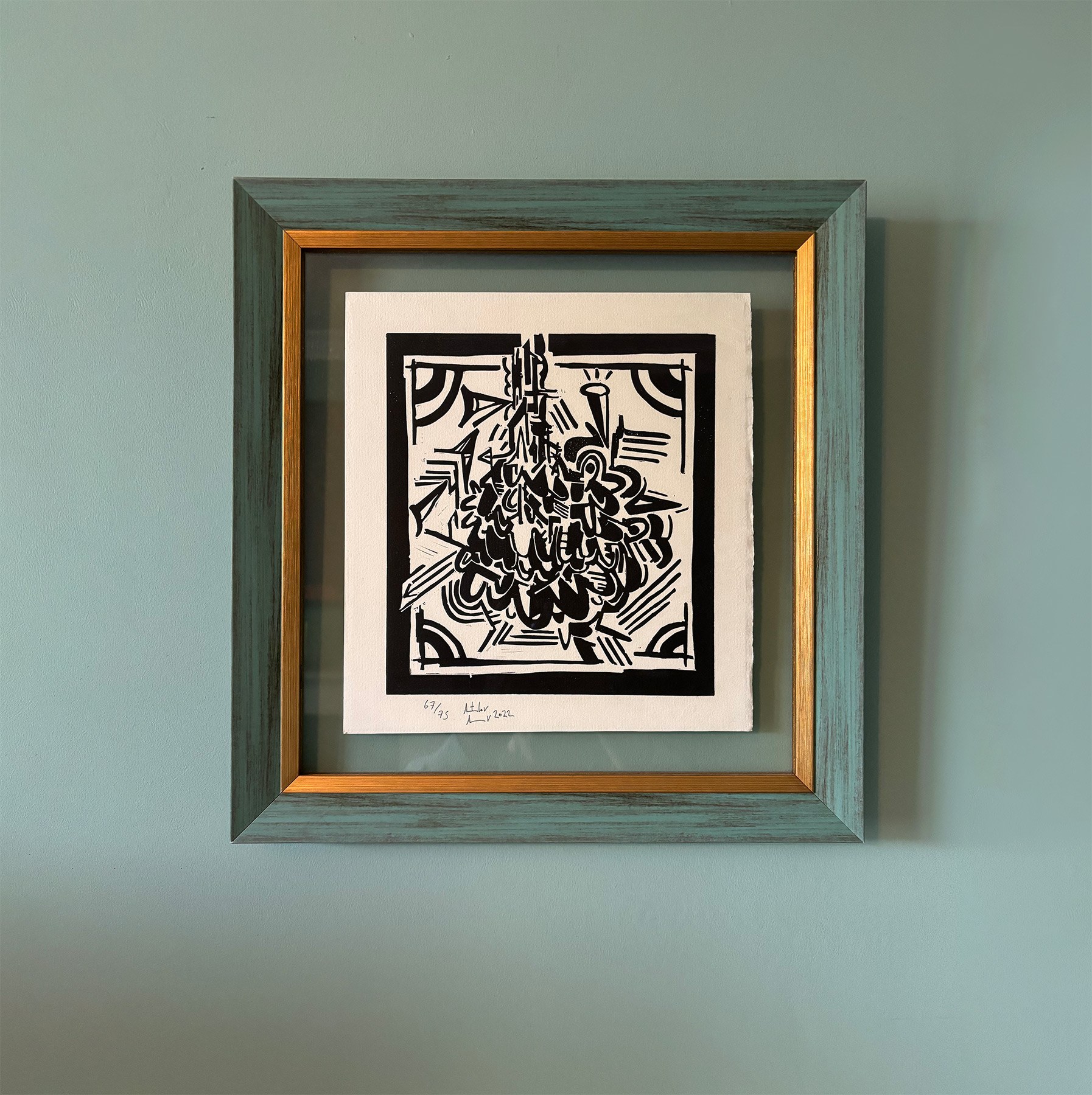 A framed black and white abstract artwork featuring bold, sharp lines and geometric shapes. The frame is textured in teal with a gold inner border. The artwork is signed and dated.