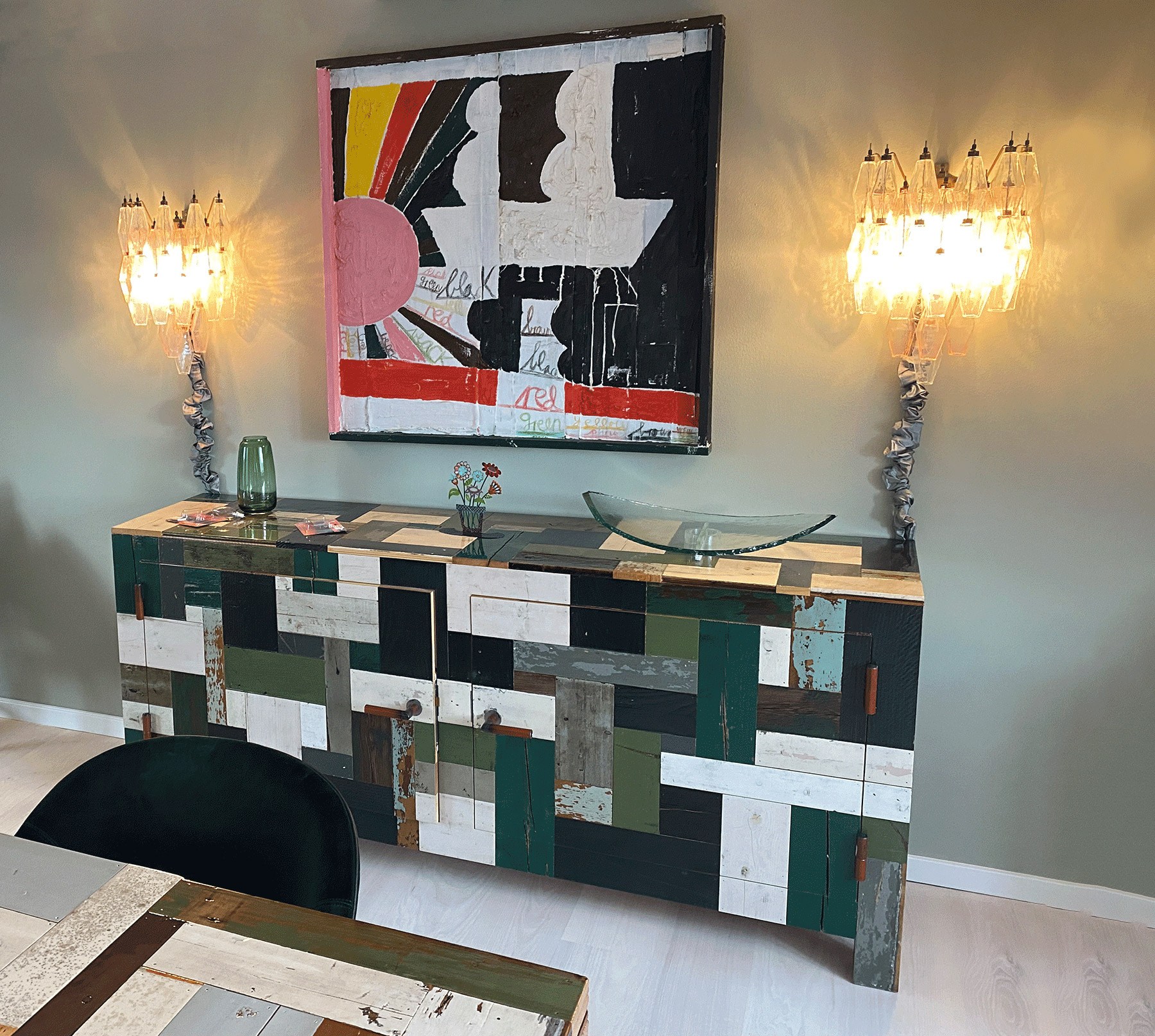 A room with a modern art painting on a green wall, flanked by two decorative wall lamps. Below is a patterned sideboard with a patchwork design of different colored panels, and a glass vase with flowers on top. A green chair is partially visible.