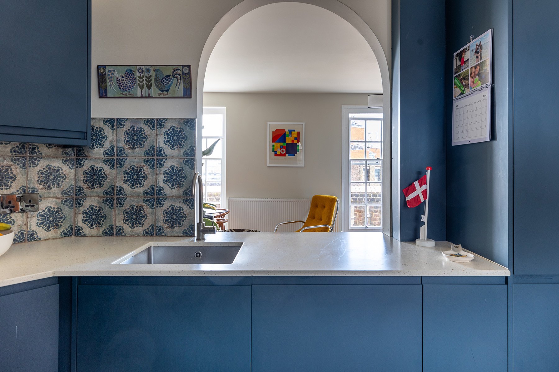 A modern kitchen with blue cabinets, a patterned tile backsplash, and a sink. An archway reveals a cozy room with a yellow armchair, colorful artwork, and bright windows. A Danish flag is displayed on the counter next to a calendar.