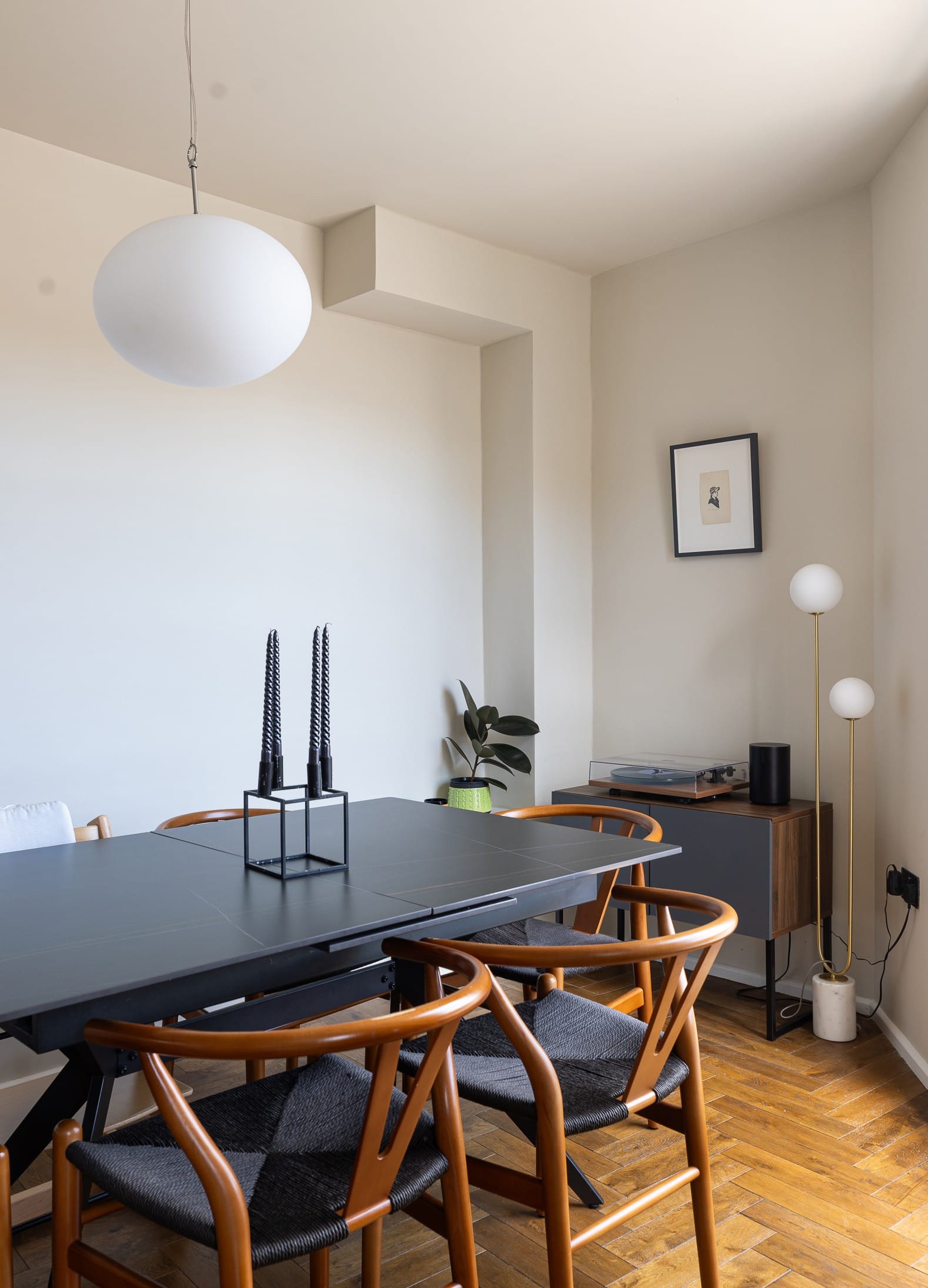 A modern dining room features a black table, wooden chairs with woven seats, and two tall black candleholders. A minimalist artwork hangs on a white wall, and a potted plant sits on a side table beside floor lamps. The flooring is wooden.