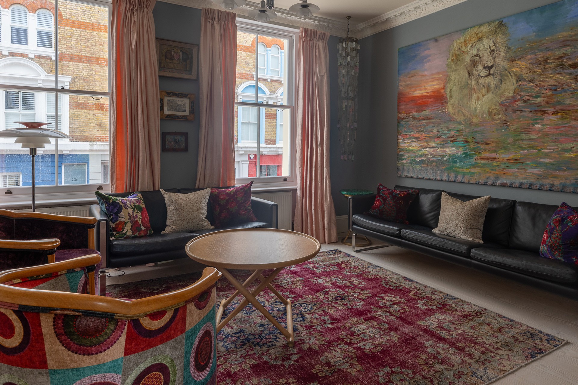 A cozy living room with two black sofas, colorful cushions, and a round wooden coffee table. The walls feature a large lion painting and several small artworks. The room is decorated with a vibrant rug and two patterned armchairs.