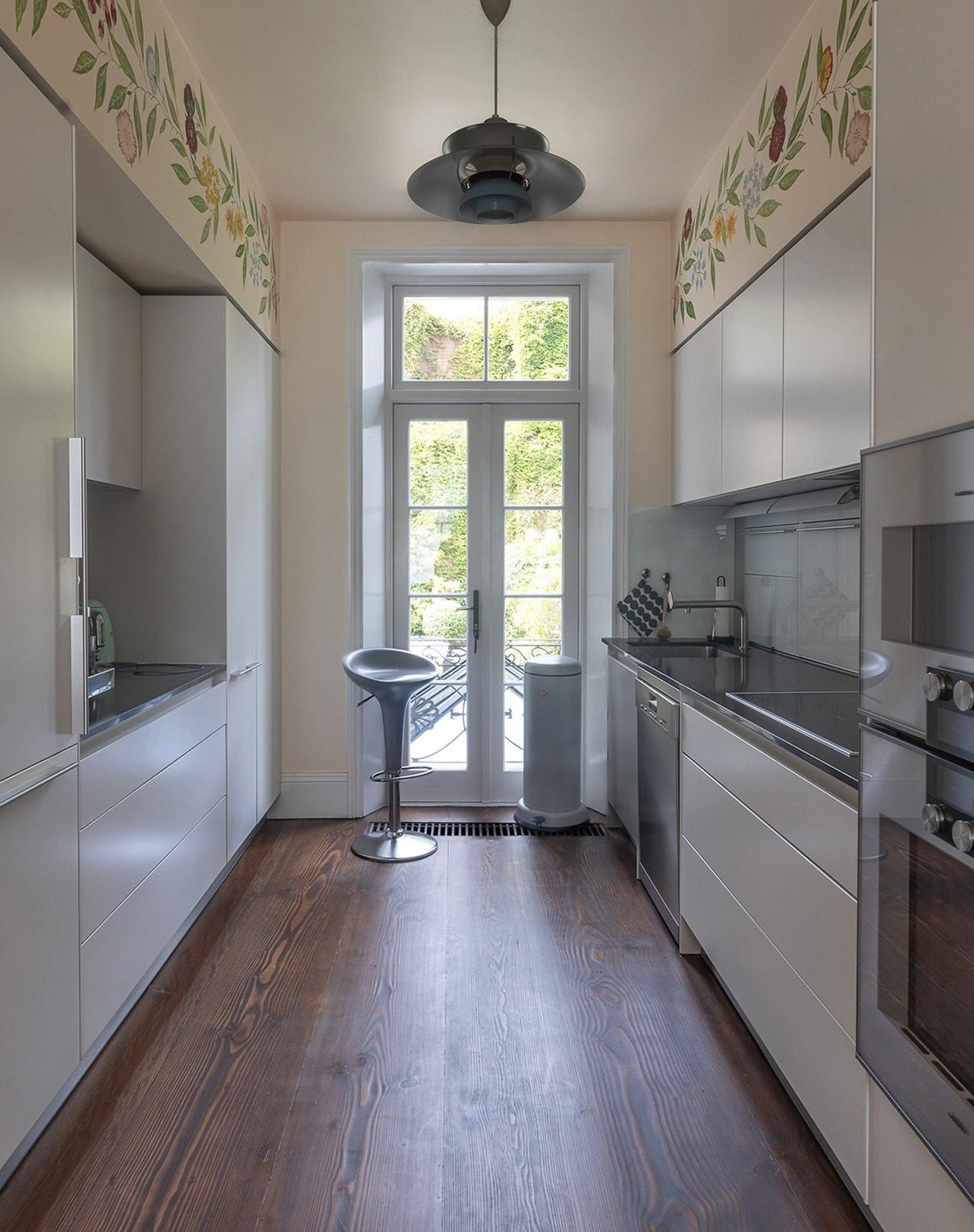 A narrow, modern kitchen with sleek gray cabinets, dark wooden floors, and a glass door leading outside. A high-backed chair sits near the door. The ceiling features floral decorations, and natural light brightens the space.