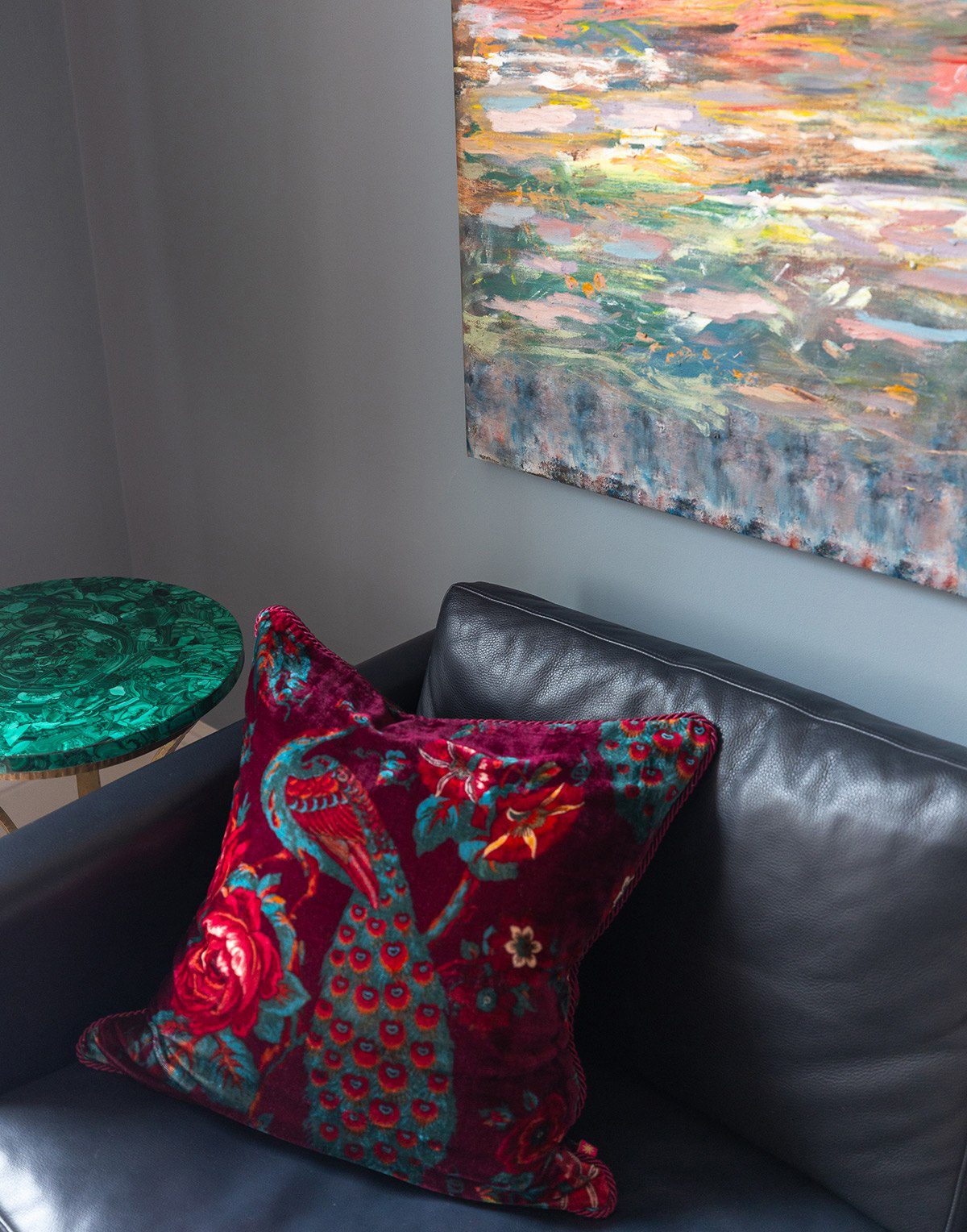 A velvet pillow with a floral and peacock design sits on a black leather chair. Behind it, a colorful abstract painting hangs on a gray wall. A round, green marble side table is partially visible beside the chair.