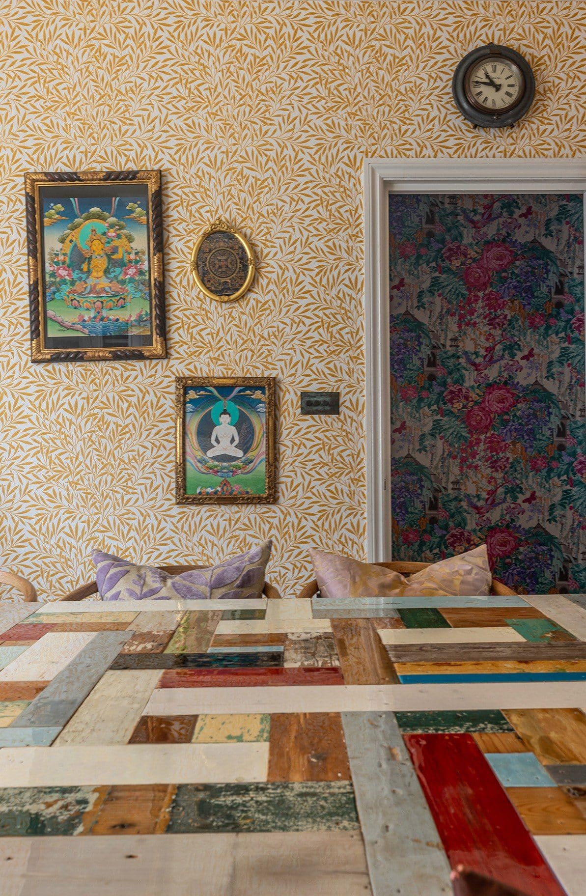 An eclectic room features a patchwork wooden table, yellow leaf-patterned wallpaper, and framed artwork on the wall. A door with floral wallpaper is partially visible. A small clock hangs above the door.