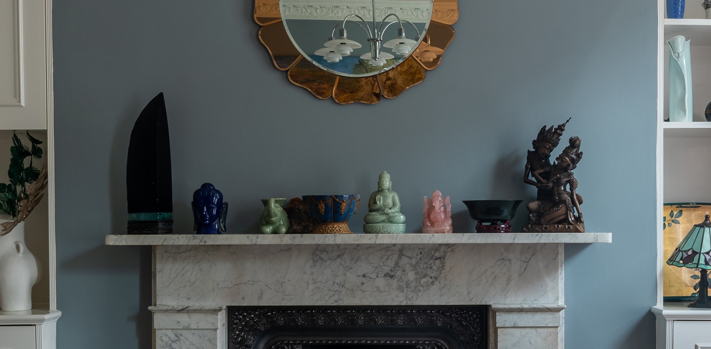 A marble fireplace mantle decorated with various statues and artifacts, including a Buddha figure, a dark sculpture, and pottery. Above is a round mirror with a decorative frame. Shelves with decorative items flank the fireplace.