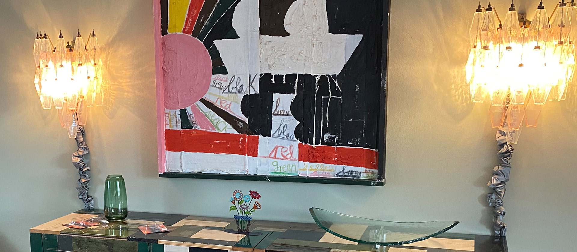 A modern art piece with abstract shapes and colors hangs between two wall sconces emitting warm light. Below, a table holds a green vase, a glass centerpiece, and decorative items in a stylish interior setting.